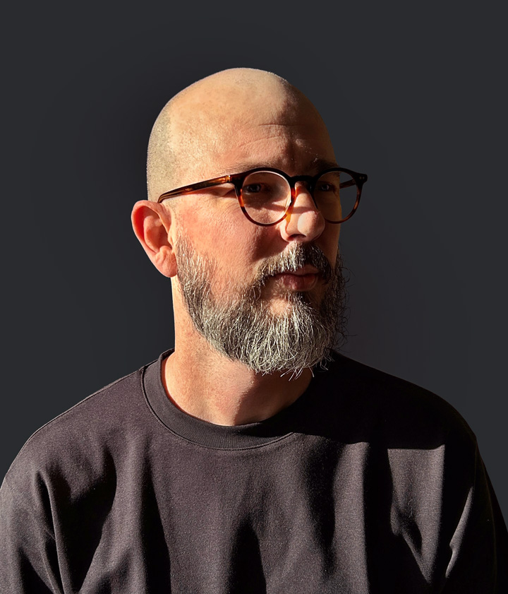 Portrait of Victor Salciotti, white man, bald, gray long beard, glasses, black t-shirt, dark gray background.